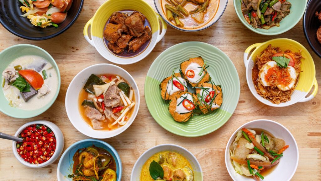 Assorted Thai food dishes including soups, curries, and stir-fries in colorful bowls on a wooden table, featuring ingredients like chili, tomatoes, and herbs.
