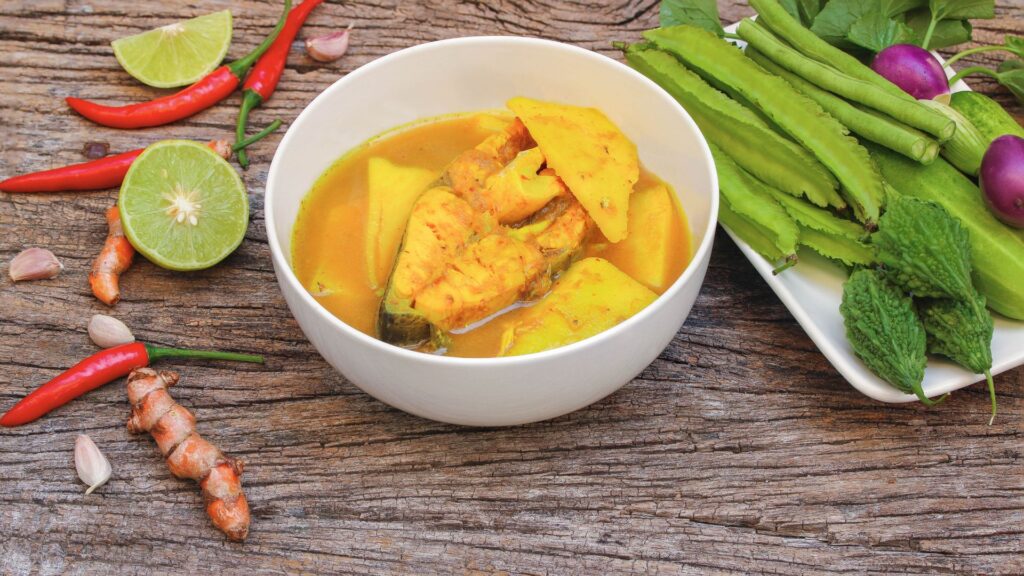 A bowl of Thai yellow curry with fish and pineapple, served on a wooden table with lime, chilies, turmeric, garlic, and a plate of green vegetables and purple eggplants.